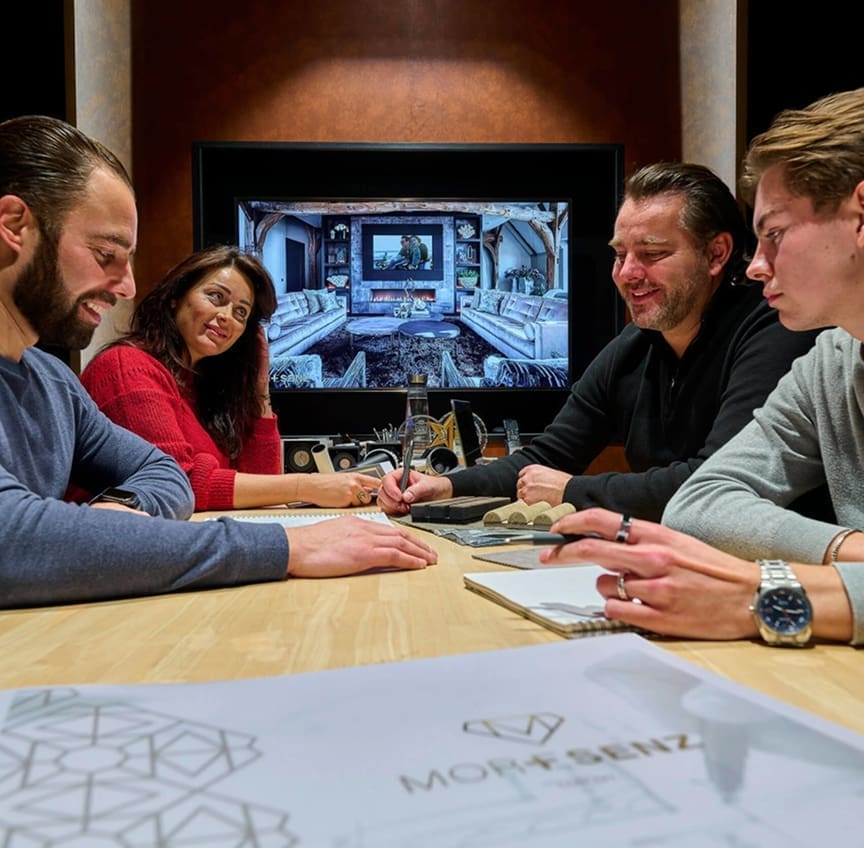 Vier mensen zitten rond een houten tafel te overleggen met op de voorgrond een bouwtekening met het Moresenz-logo. Vier mensen zitten rond een houten tafel te overleggen met op de voorgrond een bouwtekening met het Moresenz-logo.