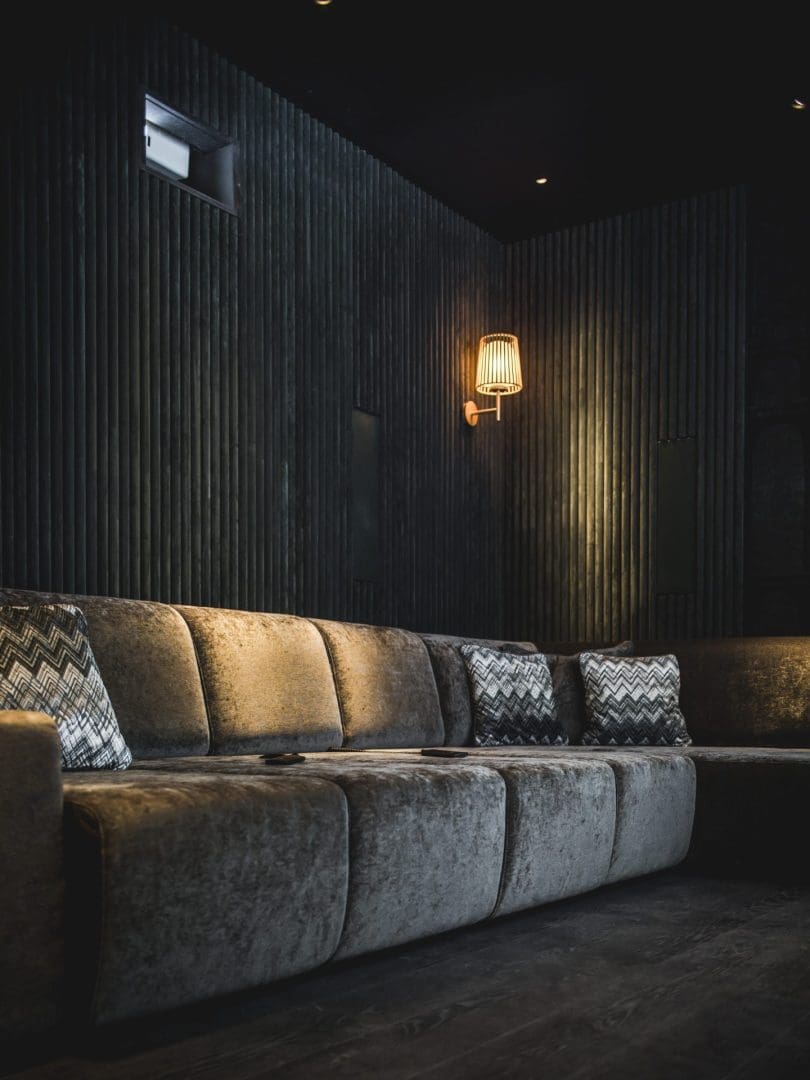 Fluwelen hoekbank met kussens tegen een donker geribbelde wand met een sfeervolle wandlamp in Project Verde 0.4. Fluwelen hoekbank met kussens tegen een donker geribbelde wand met een sfeervolle wandlamp in Project Verde 0.4.
