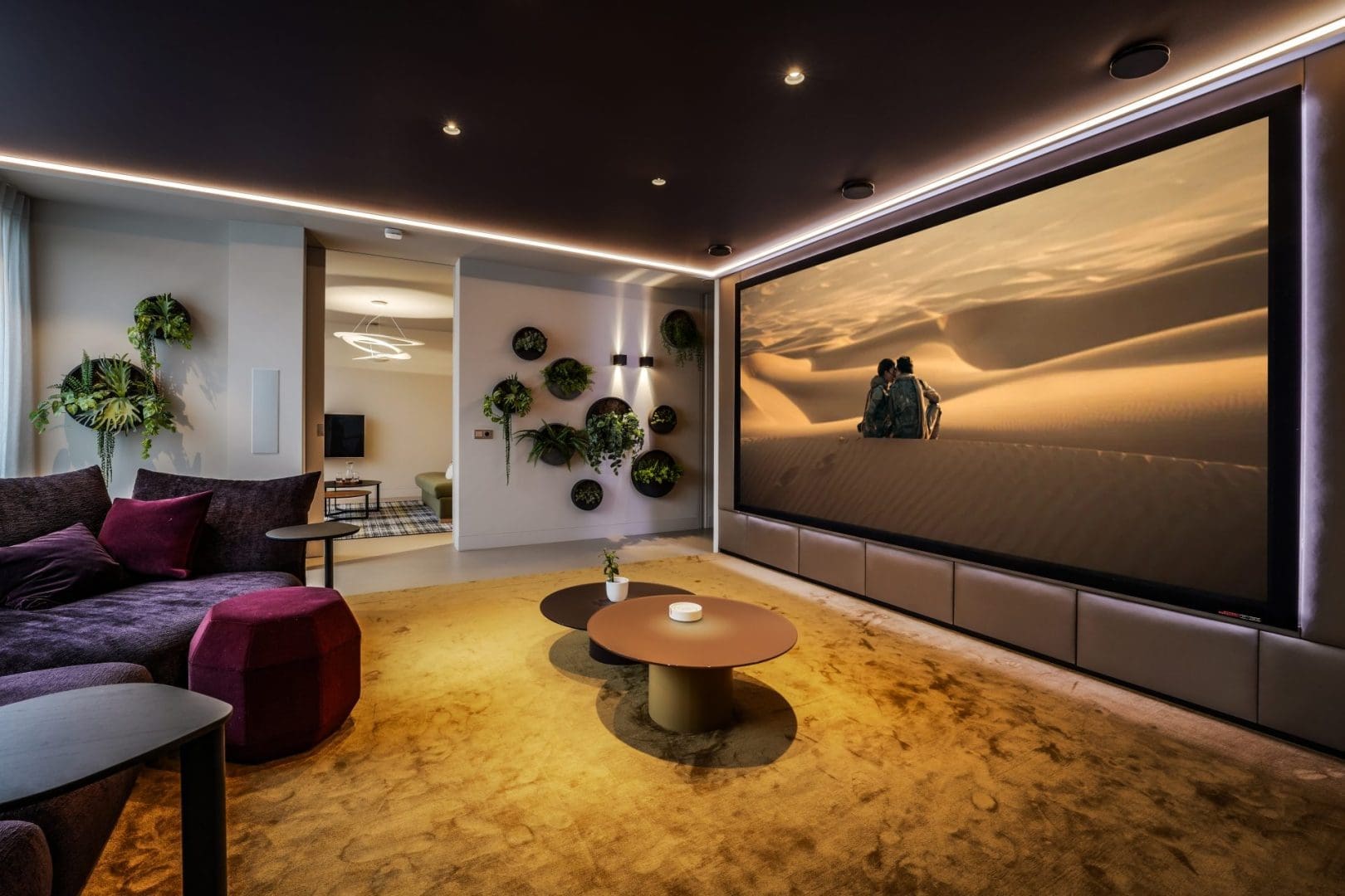 Luxe thuisbioscoop met paarse bank, gouden tapijt, ronde tafels en een groot scherm met een woestijnlandschap.