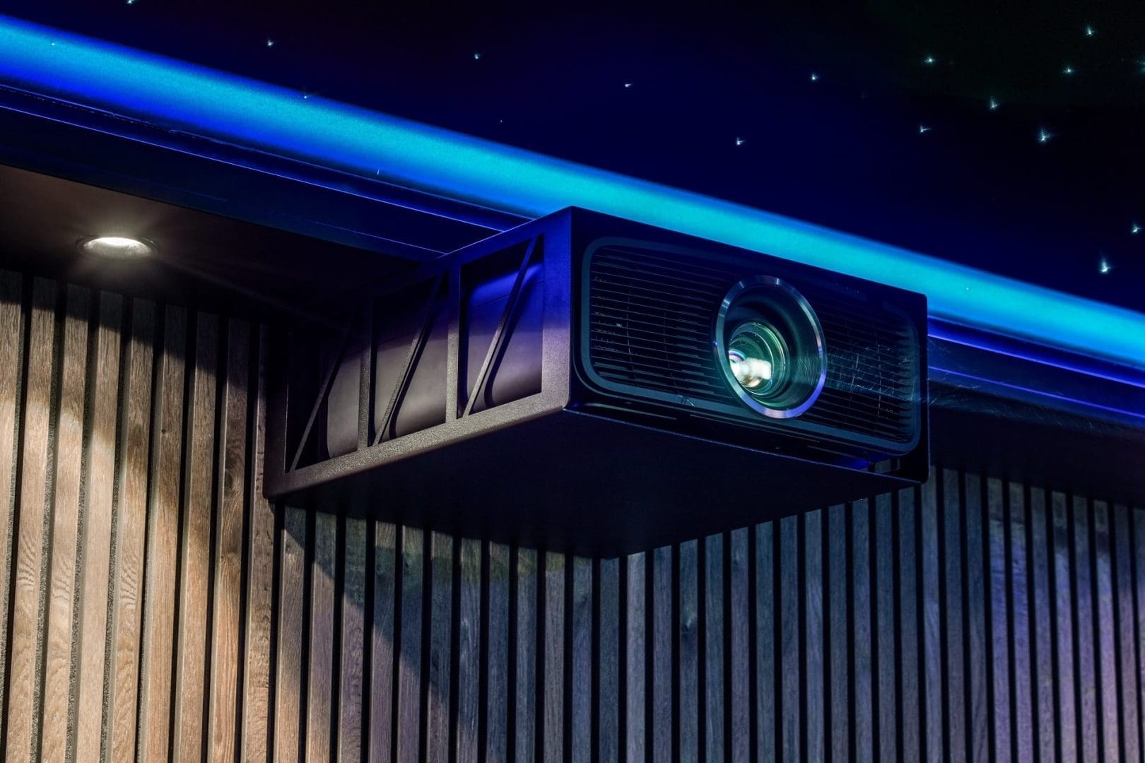 A black projector mounted on a wooden slatted wall beneath a blue-lit ceiling with a starry sky effect. A black projector mounted on a wooden slatted wall beneath a blue-lit ceiling with a starry sky effect.
