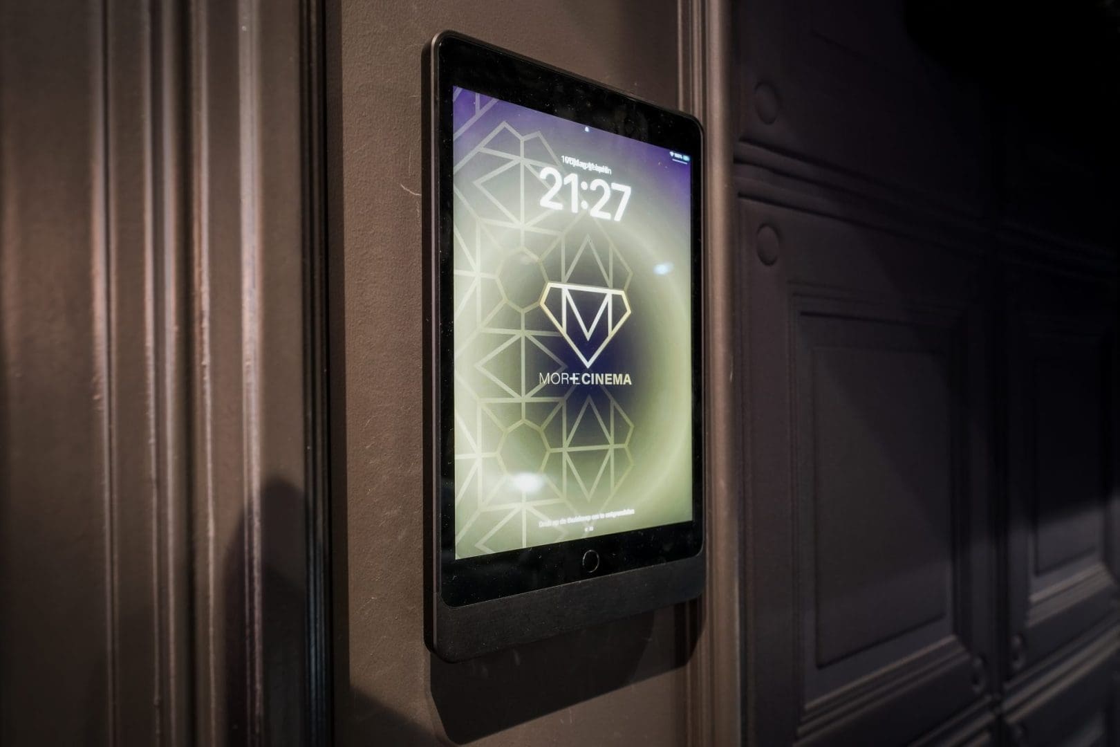 Een tablet aan een donkere muur toont de tijd 21:27 en het logo van MORE CINEMA met een geometrisch patroon. Een tablet aan een donkere muur toont de tijd 21:27 en het logo van MORE CINEMA met een geometrisch patroon.