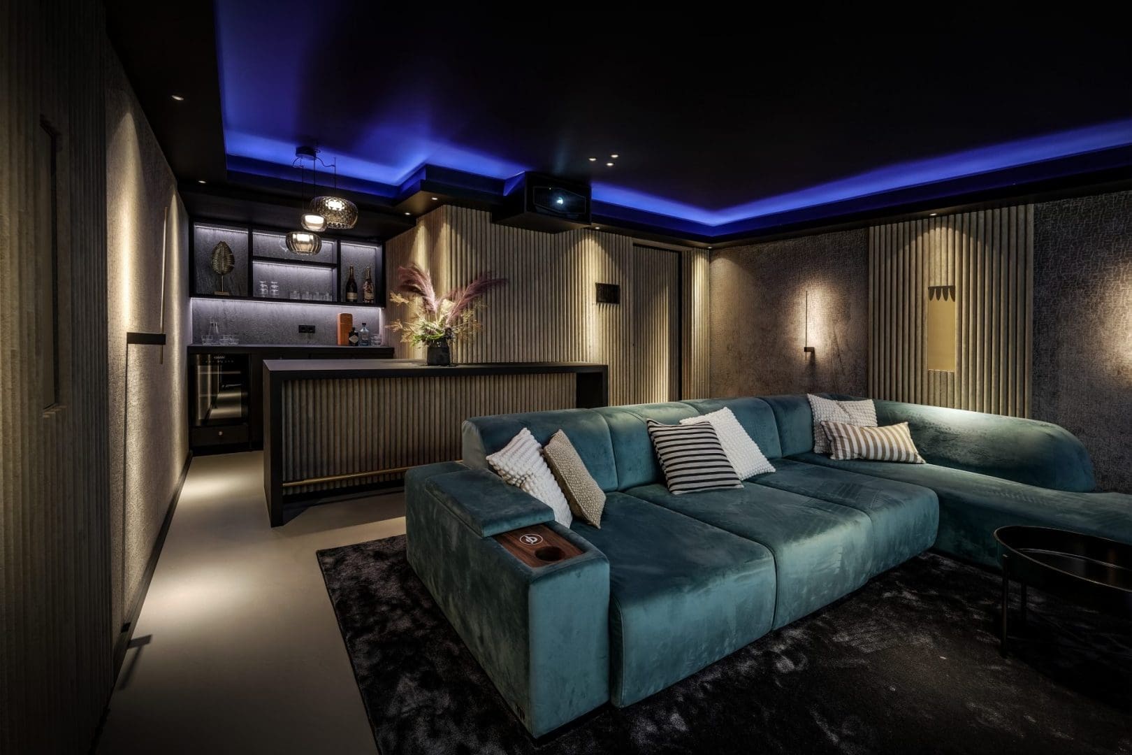 Luxe thuisbioscoop met een grote blauwe hoekbank, een bar met verlichting en sfeervolle blauwe plafondverlichting.