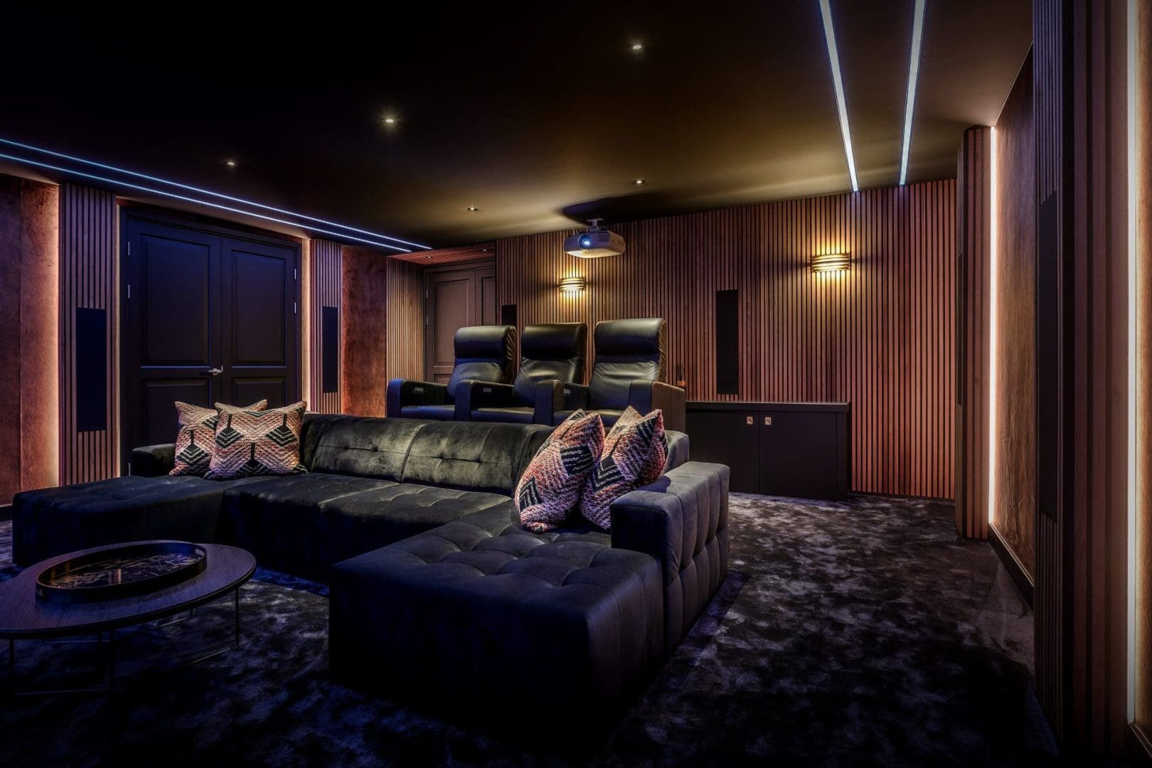 Luxe thuisbioscoop met een zwarte hoekbank, drie bioscoopstoelen, houten wandpanelen en sfeervolle ledverlichting.
