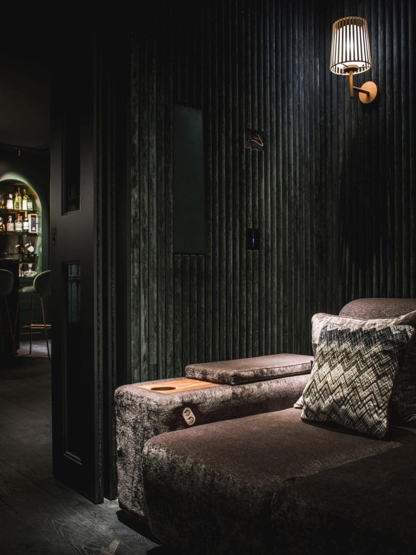 Een donkere loungehoek met een fluwelen bank tegen een geribbelde wand, verlicht door een stijlvolle wandlamp. Een donkere loungehoek met een fluwelen bank tegen een geribbelde wand, verlicht door een stijlvolle wandlamp.