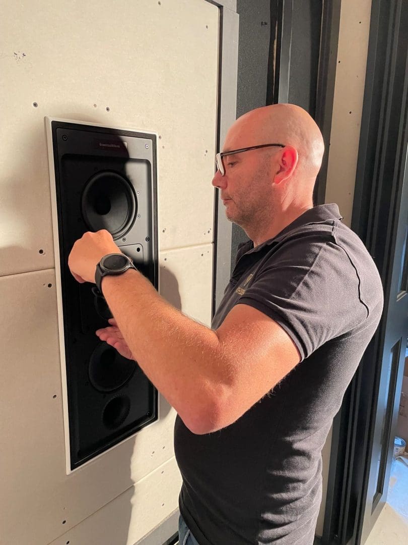 Een man met een bril installeert een Bowers & Wilkins inbouwluidspreker in een gipswand. Een man met een bril installeert een Bowers & Wilkins inbouwluidspreker in een gipswand.